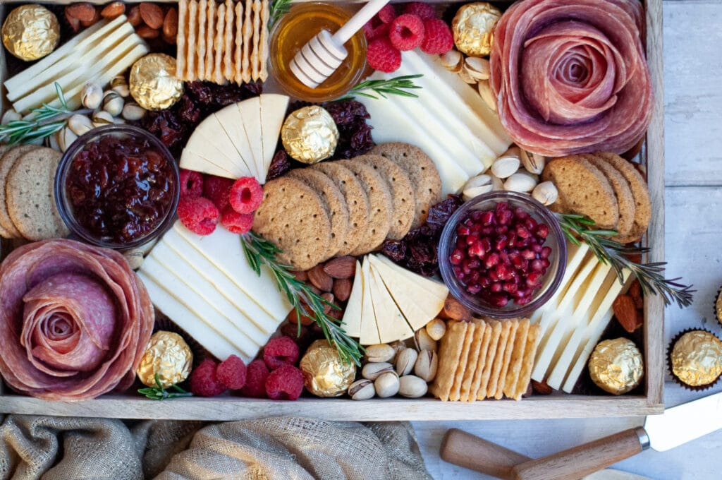 Top down view of this holiday cheese board. The board is filled with various cheeses, salami roses, crackers, nuts, fresh and dried fruits, spreads, and specialty chocolates. There are cheese knives and a golden linen next to the cheese tray.