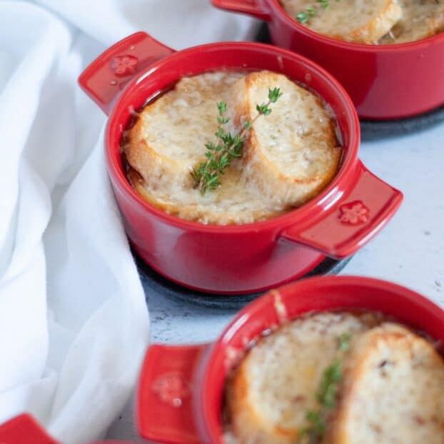 close up of a red ramiken of french onion soup, topped with french bread, cheese, and fresh thyme. Additional crocks of french onion soup surround the main dish.