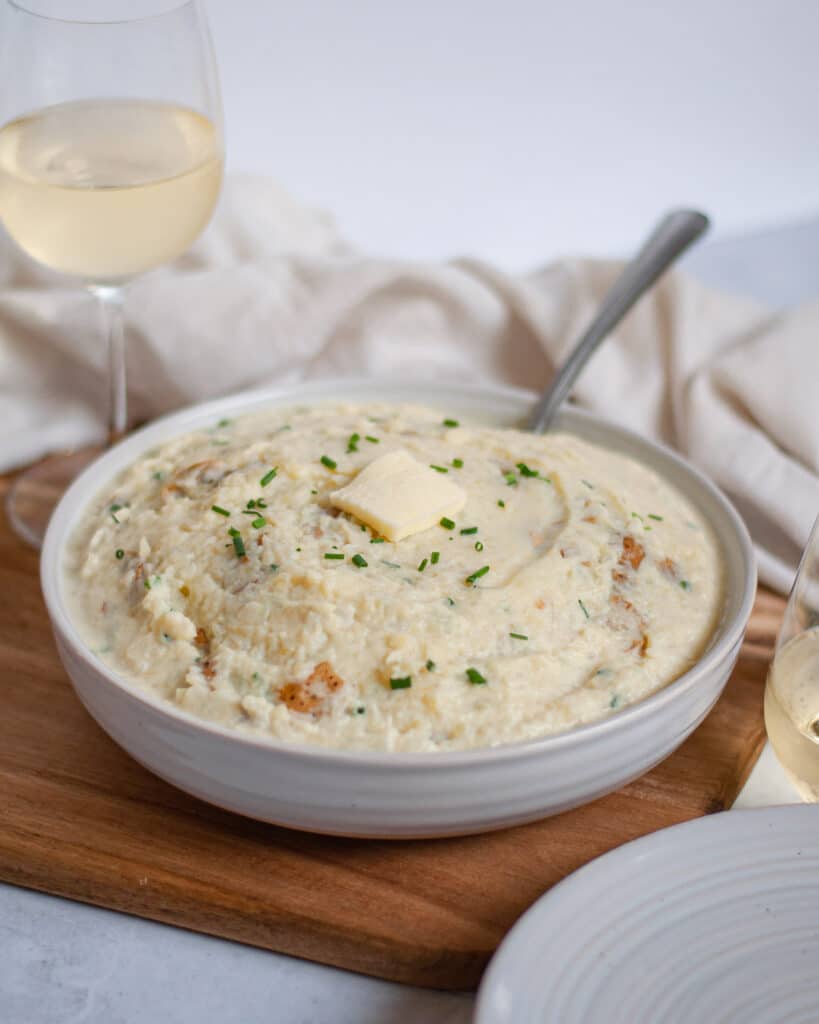 Serving bowl packed high with skinny mashed potatoes. There is a large pat of butter and chives on top of the skinny mashed potatoes, a serving spoon sticks into the dish as well. 