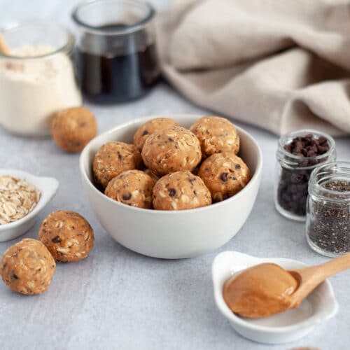 close up of a bowl of no bake peanut butter protein balls. mini chocolate chips and rolled oats are scattered around them, with a bowl filled high with additional protein bites in the background.