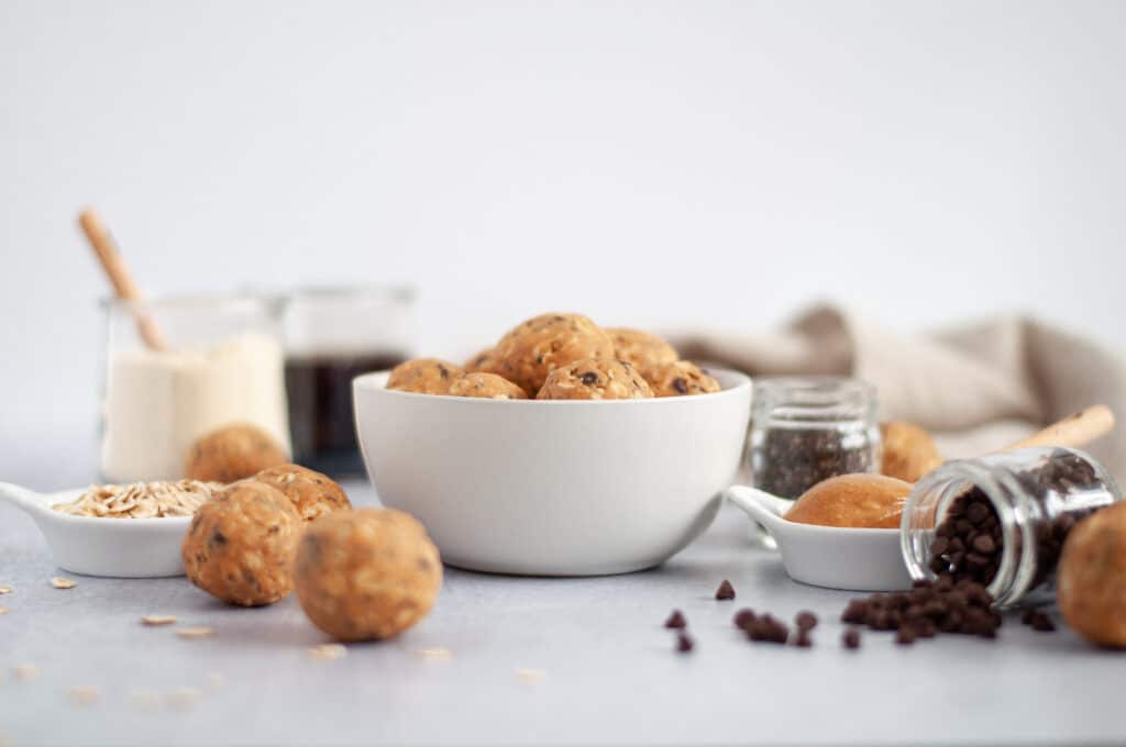 White bowl filled with peanut butter protein balls, surrounded by the ingredients that go into this protein balls recipe, including: oats, protein powder, maple syrup, chia seeds, peanut butter, and chocolate chip.