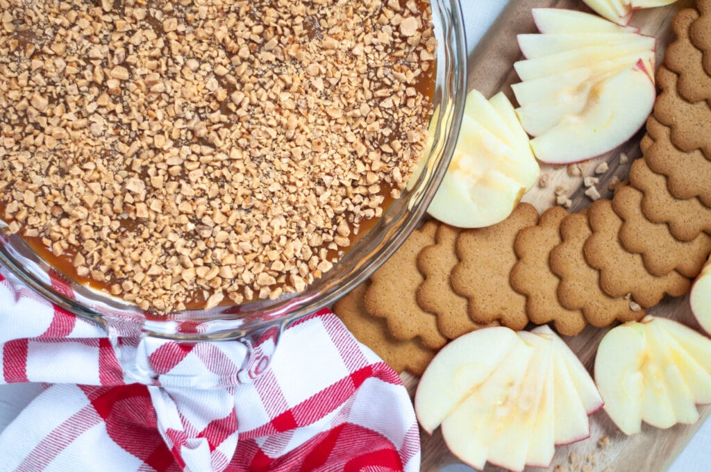 Top down view of a glass pie pan filled with this easy caramel apple dip. Next to a red and white checkered napkin and a tray of sliced apples and ginger snap cookies, ready to be dipped into this easy no bake dessert!