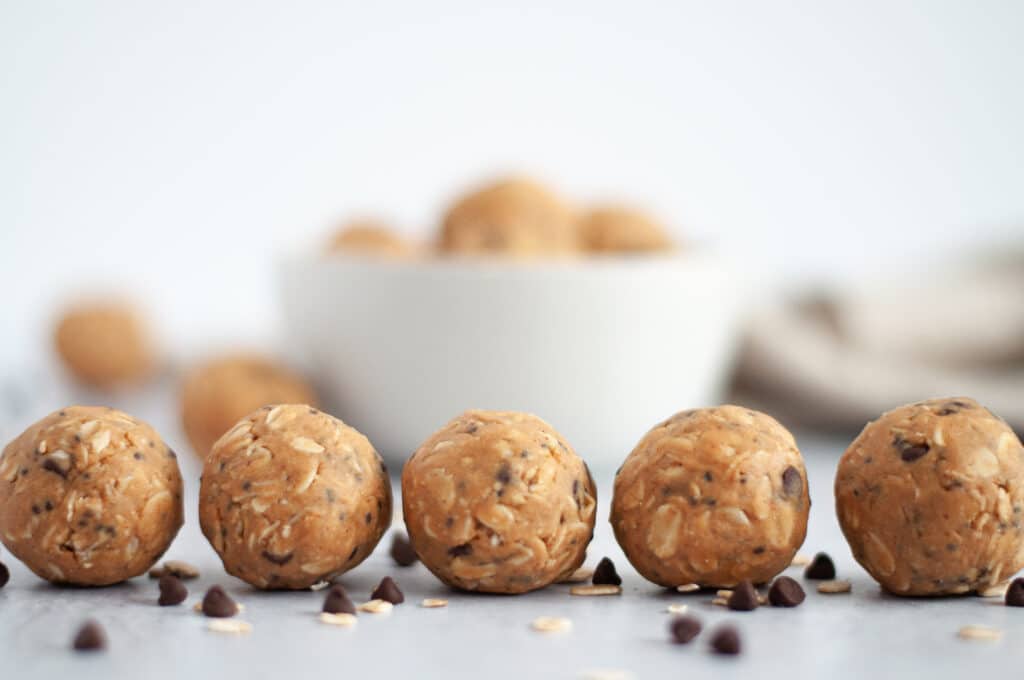 Line of peanut butter protein balls in the foreground, with a blurred out white bowl of energy bites in the background.
