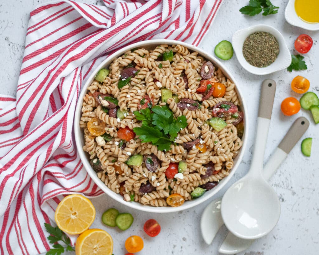 Top down view of a serving bowl filled with this vegetarian pasta salad surrounded by ingredients, serving spoons, and a striped napkin.