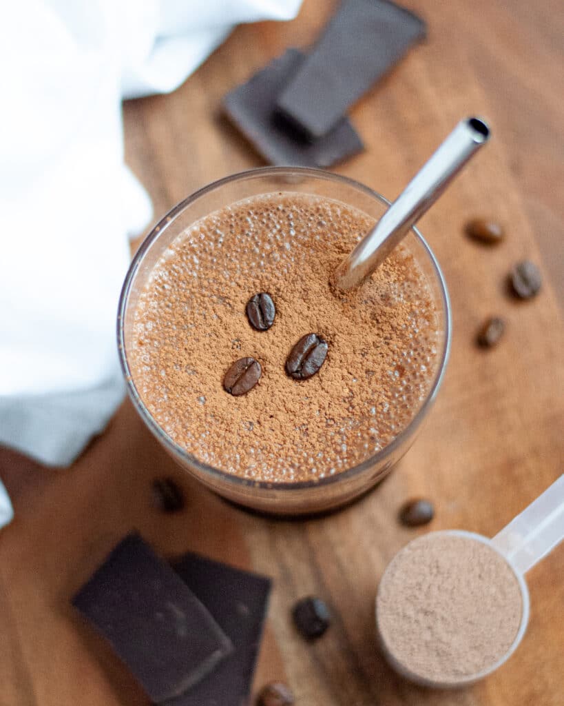 A glass filled with this coffee chocolate protein smoothie recipe, dusted with cocoa powder and topped with three coffee beans for decoration. The glass has a metal straw, and is surrounded by chocolate pieces, coffee beans, a scoop of chocolate protein powder, and a white linen.
