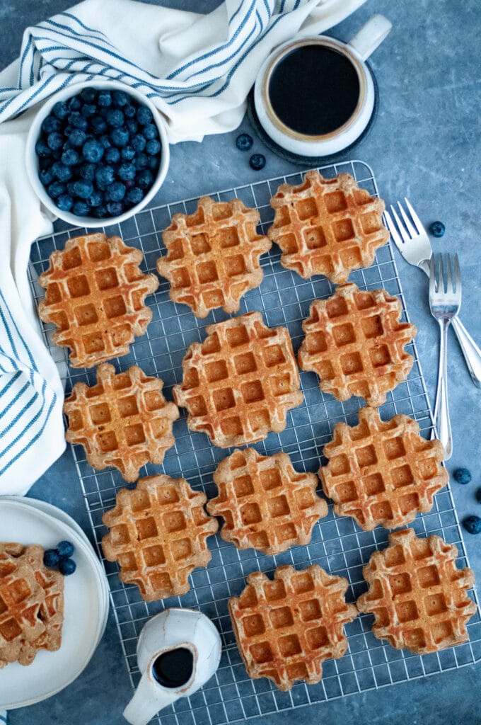 Top down view of this healthy waffle recipe displayed on a wire cooling rack with a carafe of maple syrup, bowl of blueberries, and a cup of coffee