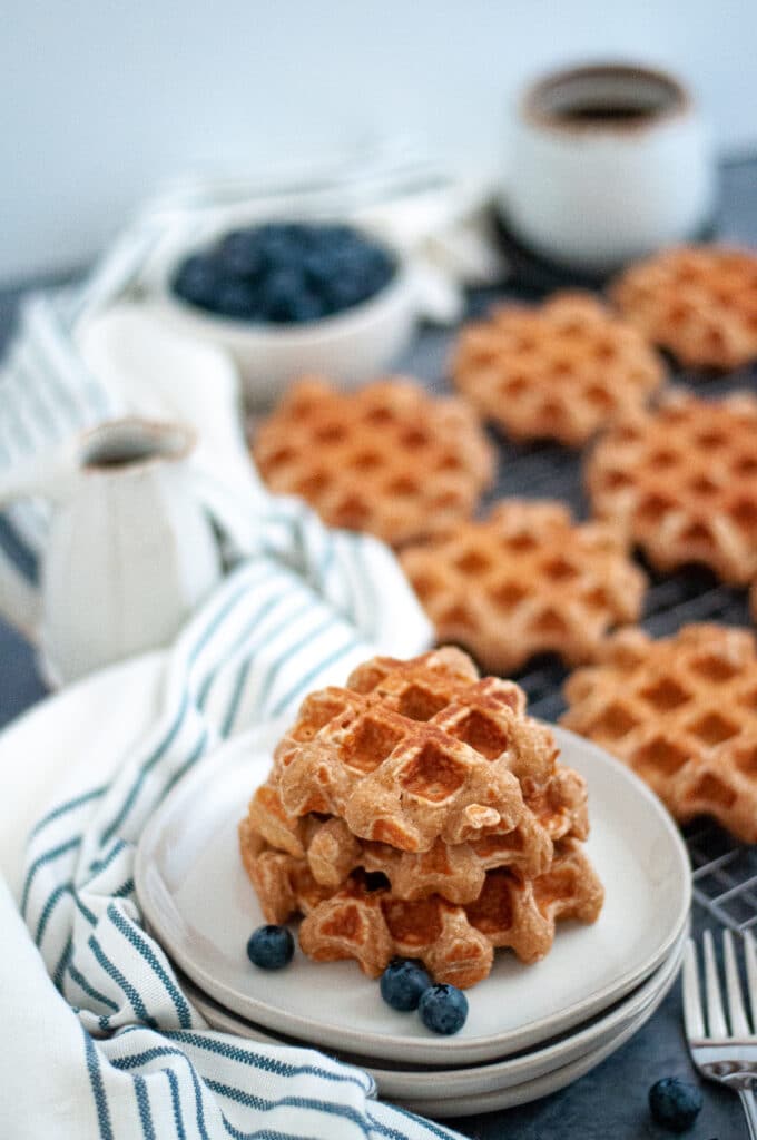 A stack of homemade waffles on a plate with additional whole wheat waffles on a cooling rack in the background