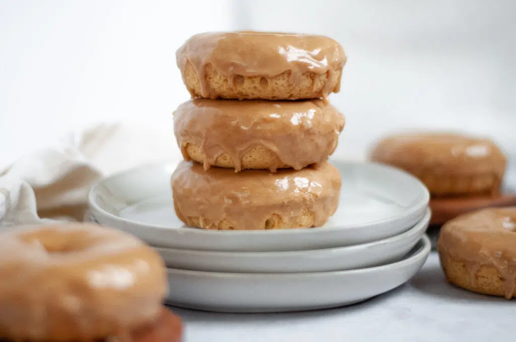 Three glazed donuts are stacked on a white plate with another donut partially visible behind the stack. Additional glazed donuts are in the background, slightly out of focus. The setting is minimalistic with soft lighting.