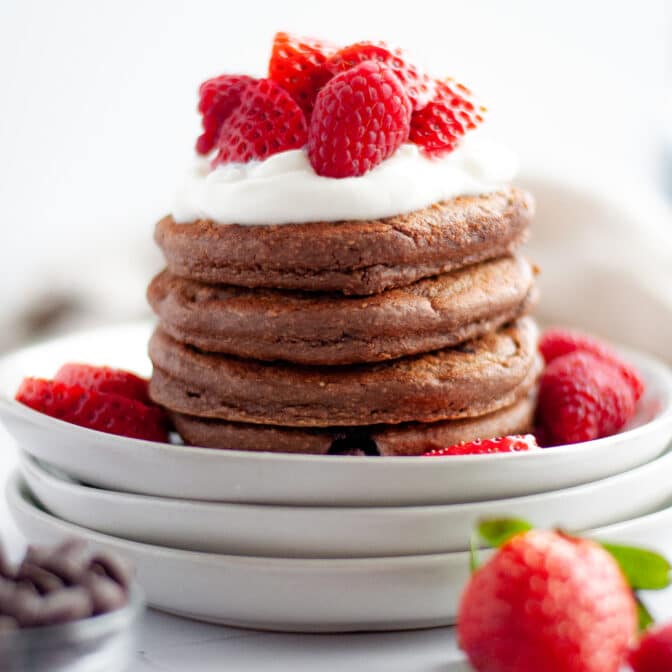 a stack of chocolate protein pancakes topped with yogurt and strawberries on a stack of white plates. the plate is surrounded by more strawberries, a small bowl of chocolate chips, and a scoop of chocolate protein powder.