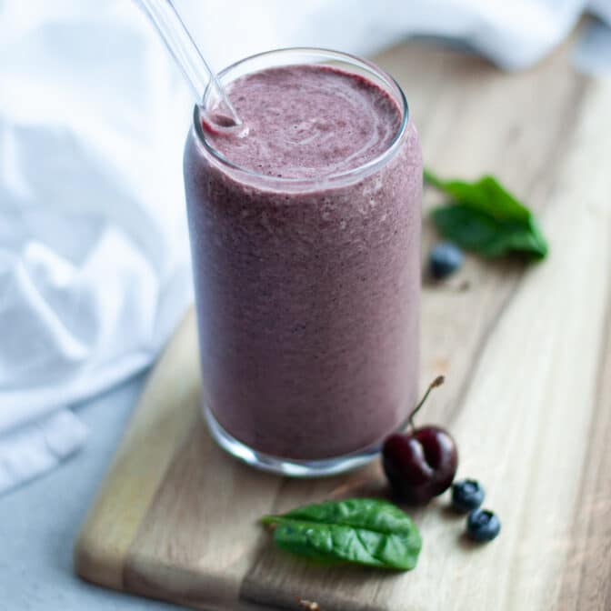close up for of a cherry berry smoothie. The glass is sitting on a wooden board with cherries, blueberries, and fresh spinach leaves around it.