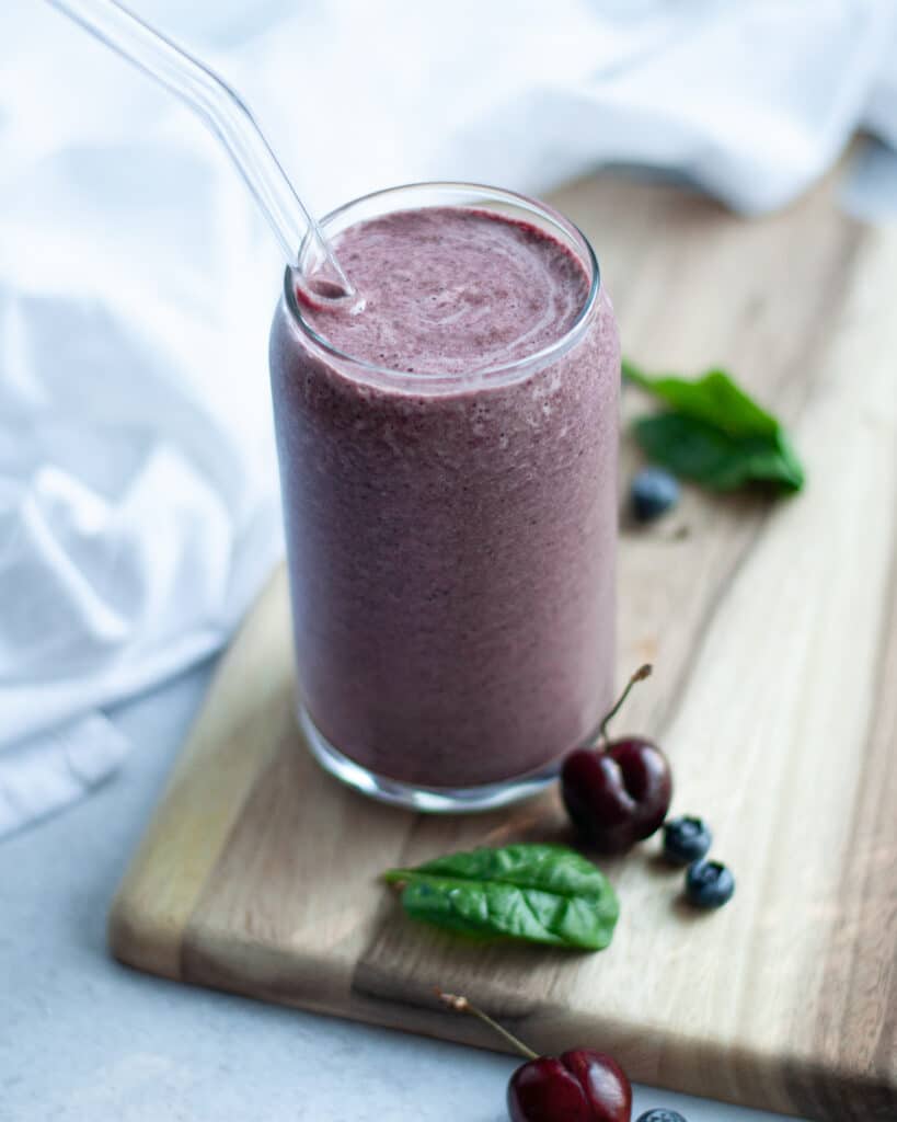 Glass of this cherry berry smoothie with coconut water sitting on a wood board with a glass straw. This healthy smoothie is surrounded by ingredients and a napkin.