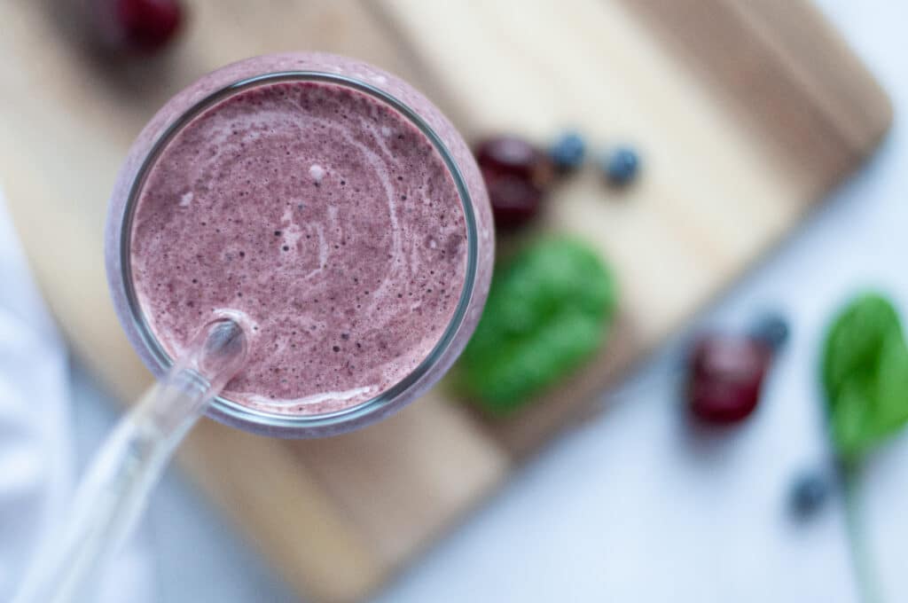 Top down view of a glass filled with this cherry and blueberry protein shake recipe including a glass straw. The glass is sitting on a wooden cutting board and is surrounded bycherries, blueberries, and spinach.