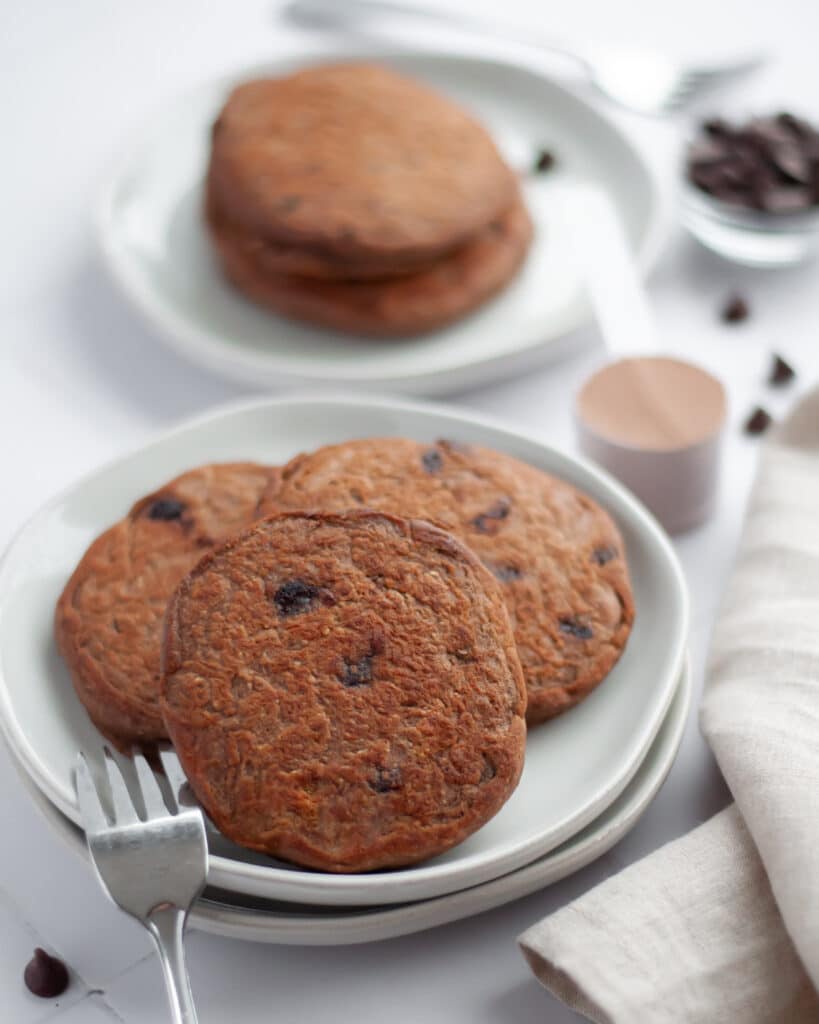 A plate filled with chocolate chip pancakes ready for someone to dig in. There is a scoop of protein powder, chocolate chips, a linen, and another plate with pancakes.