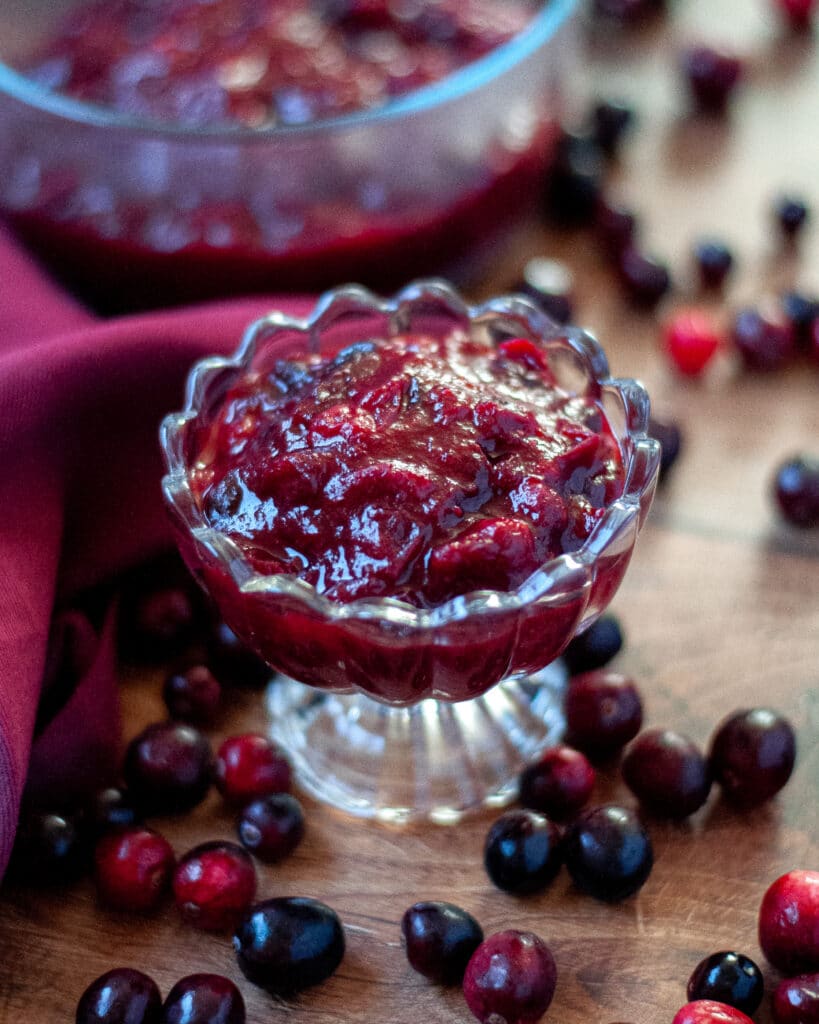 Glass dish of cranberry sauce, surrounded by fresh cranberries a dark red napkin, and a larger serving dish filled with cranberry sauce.