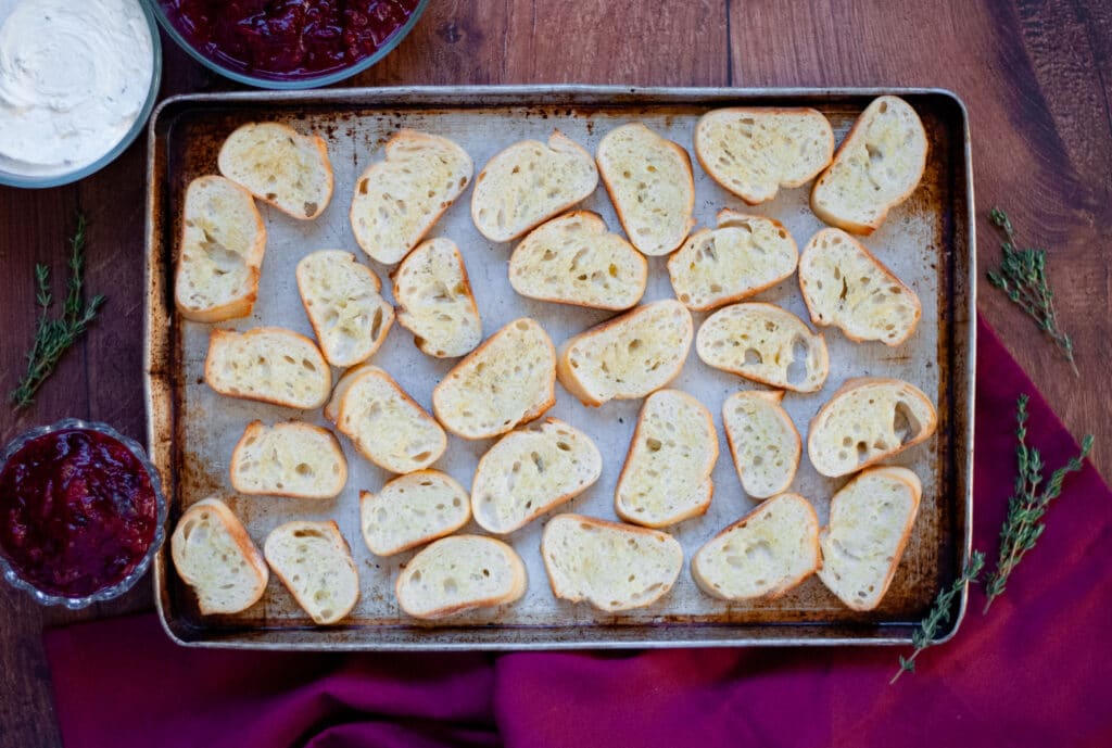 Top down view of a baking sheet full of toasted crostini, ready to be topped. Bowls of whipped goat cheese and fresh cranberries sauce, and sprigs of thyme surround the baking sheet.
