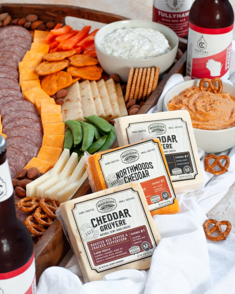 Close up of the cheeses used for this cheese board, with the full meat and cheese tray behind it, the bowl of hummus next to it, pretzels scattered around, and bottles of beer all ready to watch the big game!