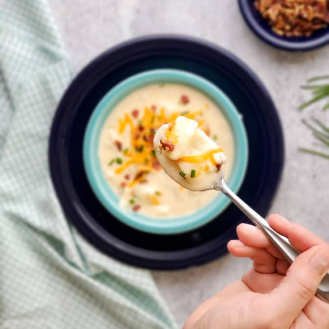 a hand holding a spoonful of cheesy potato soup above a serving bowl of soup