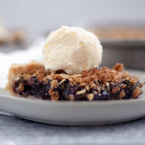 close up of a slice of blueberry slab pie with a scoop of vanilla ice cream on top. in the background you can see the pan of blueberry pie bars and another slice of slab pie.