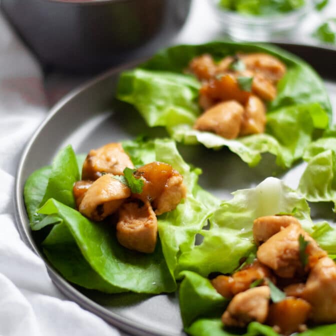 A dark grey plate filled with mango chicken lettuce wraps garnished with cilantro. There is a white linen on the side of the plate, and bowls on ingredients and additional filling in the background.