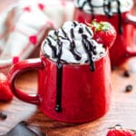 Close-up shot of two Chocolate Covered Strawberry Coffees in red mugs with whipped cream, chocolate syrup, and a strawberry on top.