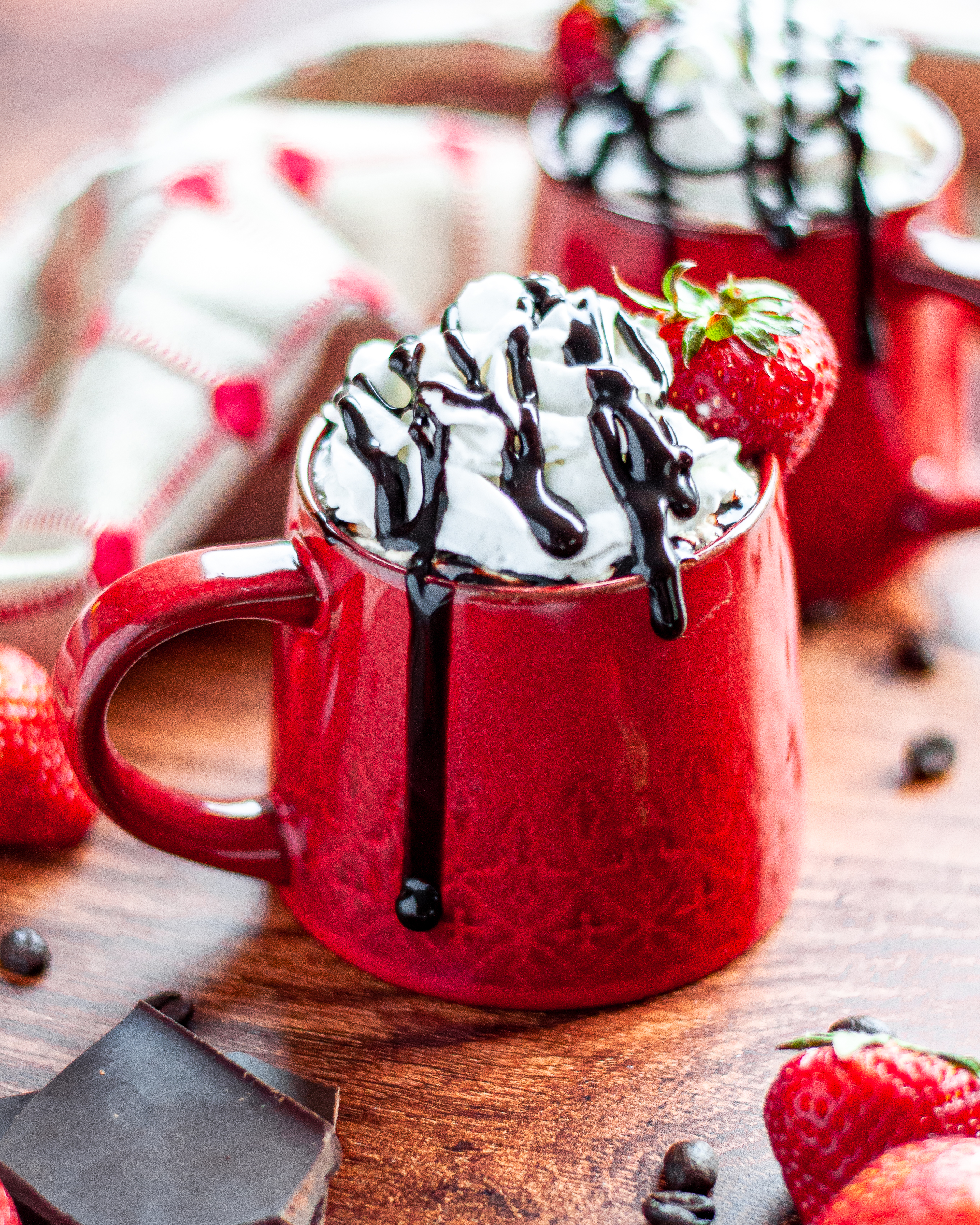 Close up of a red mug filled with a chocolate strawberry coffee and topped with whipped cream and dripping chocolate sauce. The mug is garnished with a fresh strawberry. The strawberry mocha is surrounded by pieces of chocolate, coffee beans, fresh strawberries, and tan and red linen, with another mug in the background.