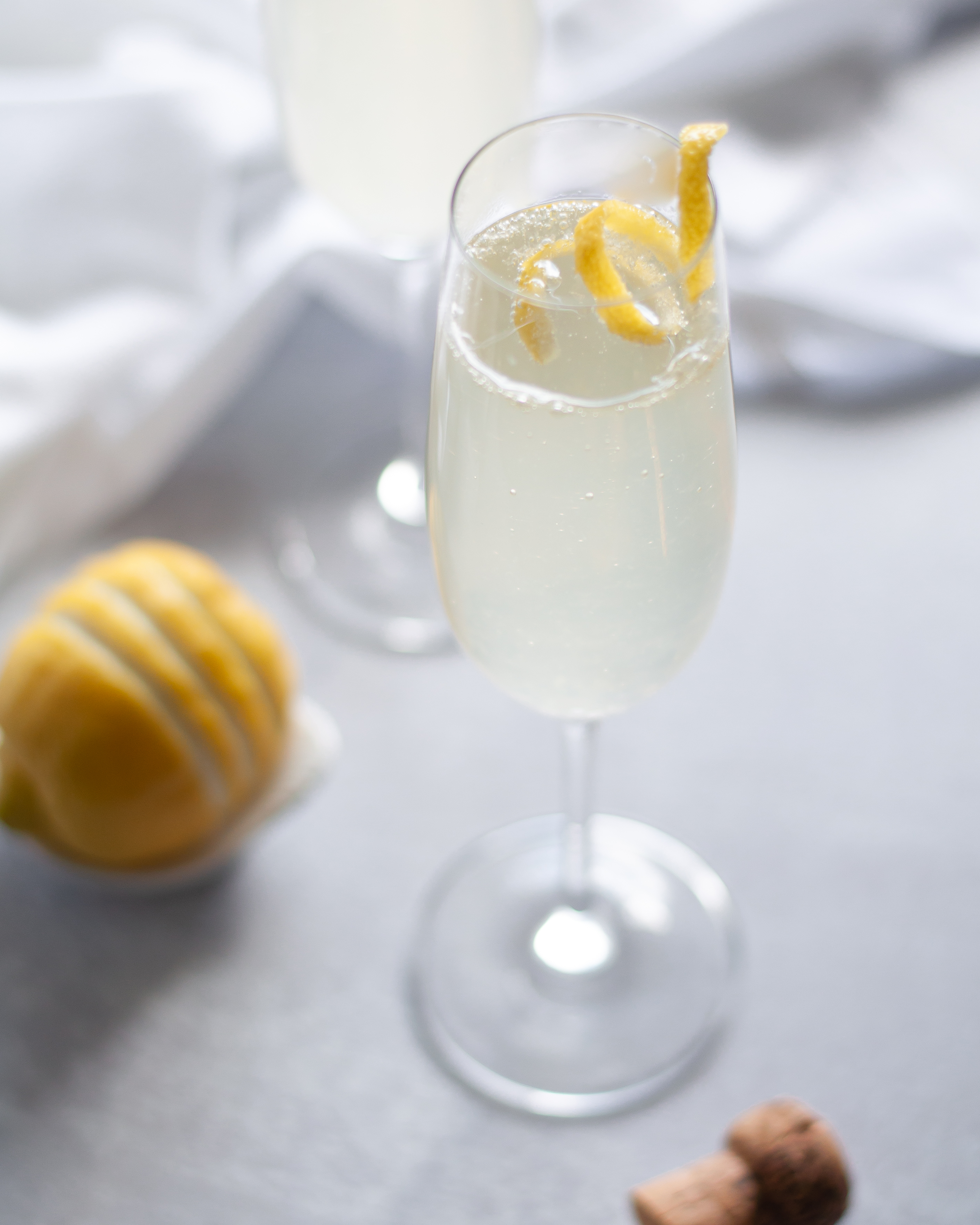 Champagne flute filled with this French 75 recipe and garnished with a lemon twist. Around the glass is a champagne cork, lemon, a 2nd cocktail, and a white napkin.