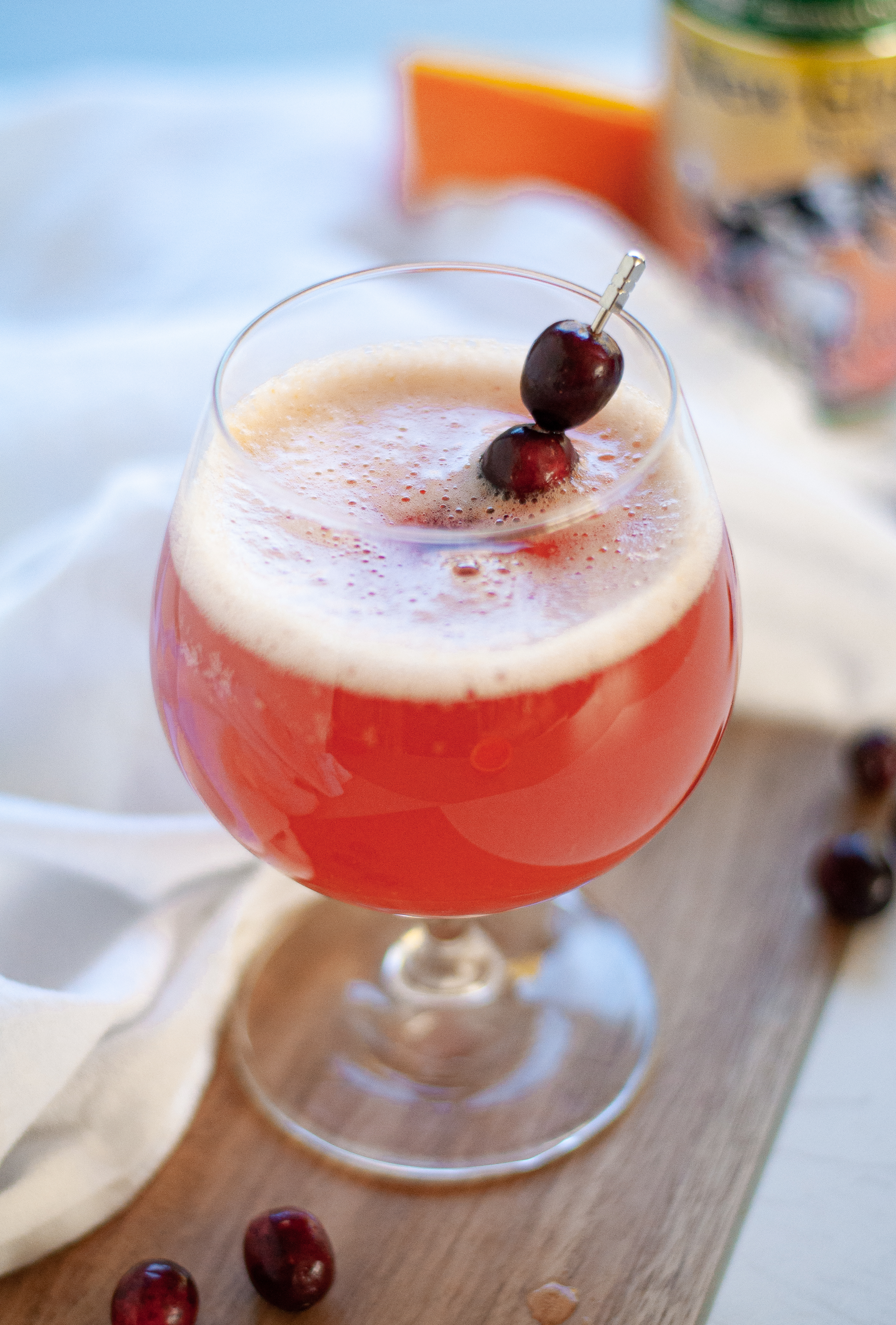 Close up of a cranberry beer shandy garnished with fresh cranberries. The glass of cranberry shandy is surrounded by fresh cranberries, a white napkin, and is in front of a can of farmhouse ale and orange halve.