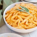 Serving bowl filled with pumpkin and boursin cheese pasta with fresh sage on top. A plate of this pasta with pumpkin sauce is peaking into the front corner of the image. There is a green napkin and additional plates in the background.