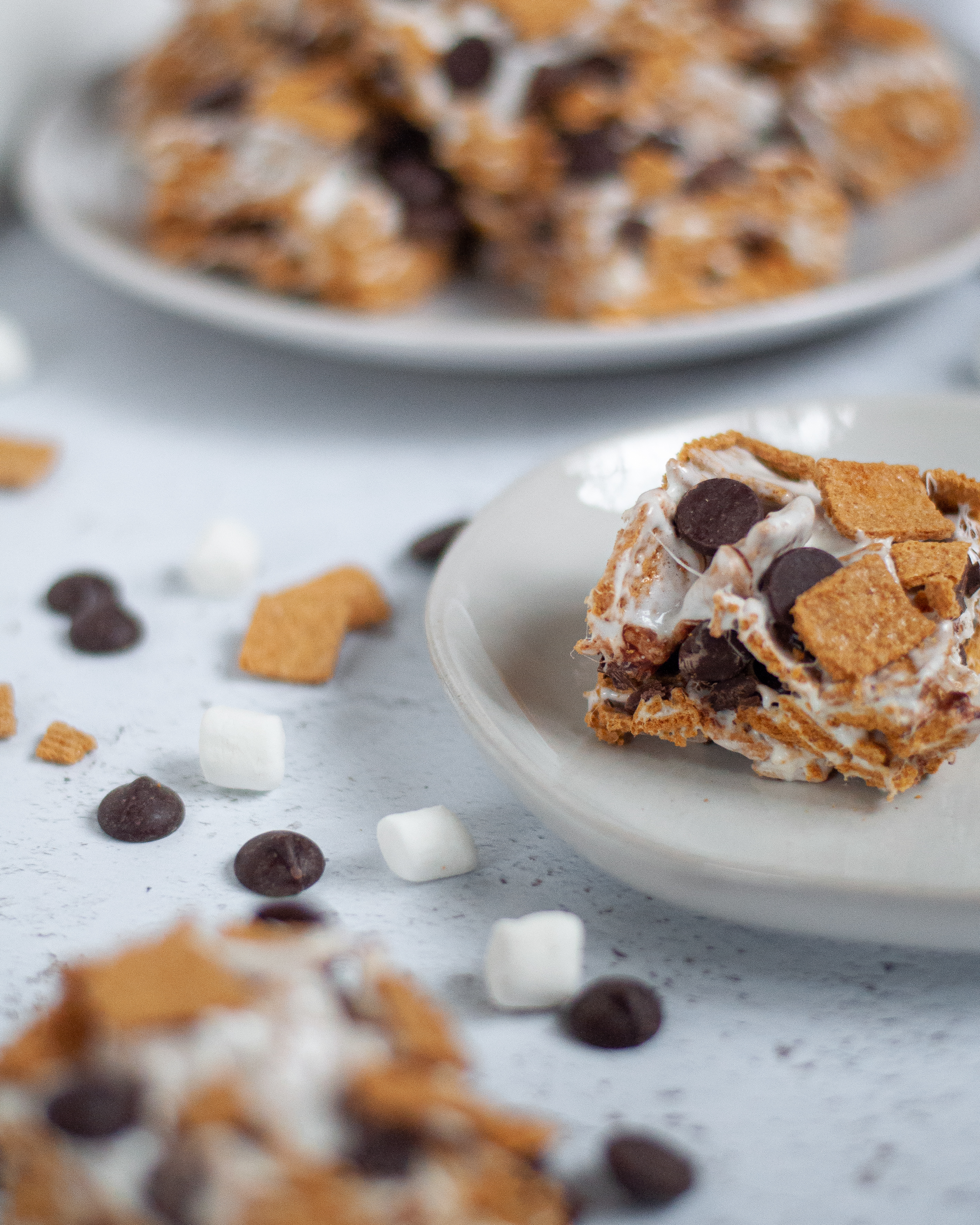 Focus on a plate with a smores bar at the center right. In the foreground there is another smores dessert bar blurred out, and in the background a platter for smores bars are also blurred. There are mini marshmallows, chocolate chips, and golden grahams scattered throughout the image as well.