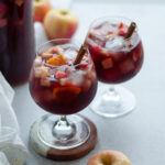 close up shot of two wine glasses filled with fall red wine sangria and garnished with a cinnamon stick. the pitcher of sangria is in the background, with apples and a white linen.