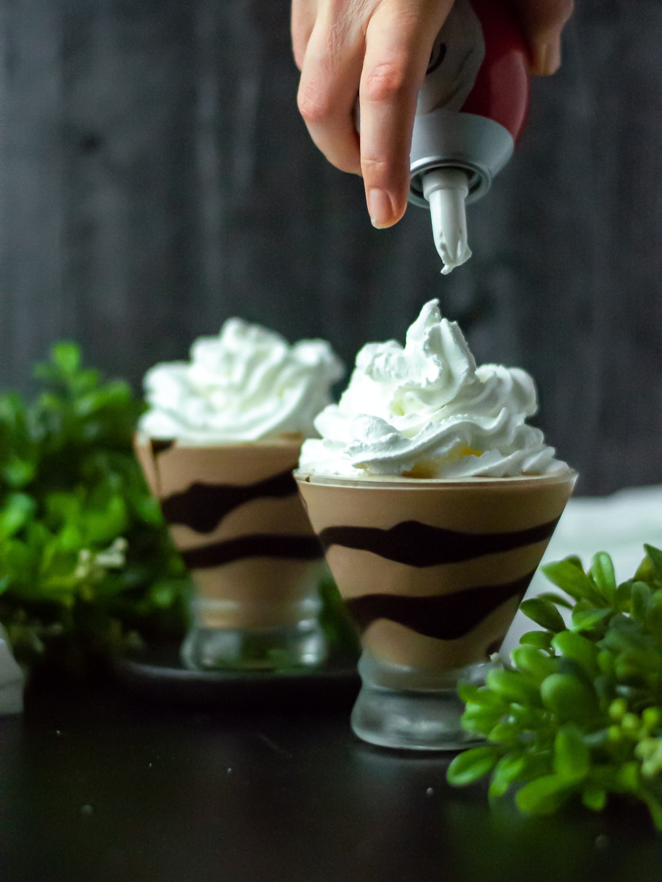 Two glasses with chocolate swirls filled with frozen mudslides and in the process of being topped with squirt whipped cream.