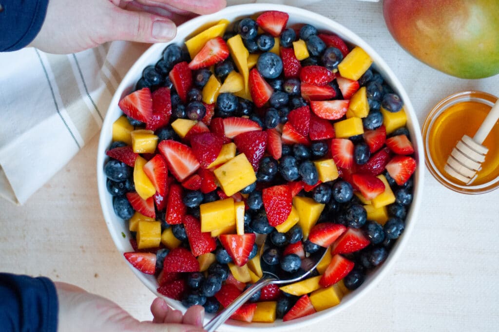 Top down view of a serving bowl filled with this healthy fruit salad recipe. Two hands are in the image, one is holding the bowl while the other is stirring the fruit salad. There is also a striped linen in the image, a bowl of honey, and a fresh mango.