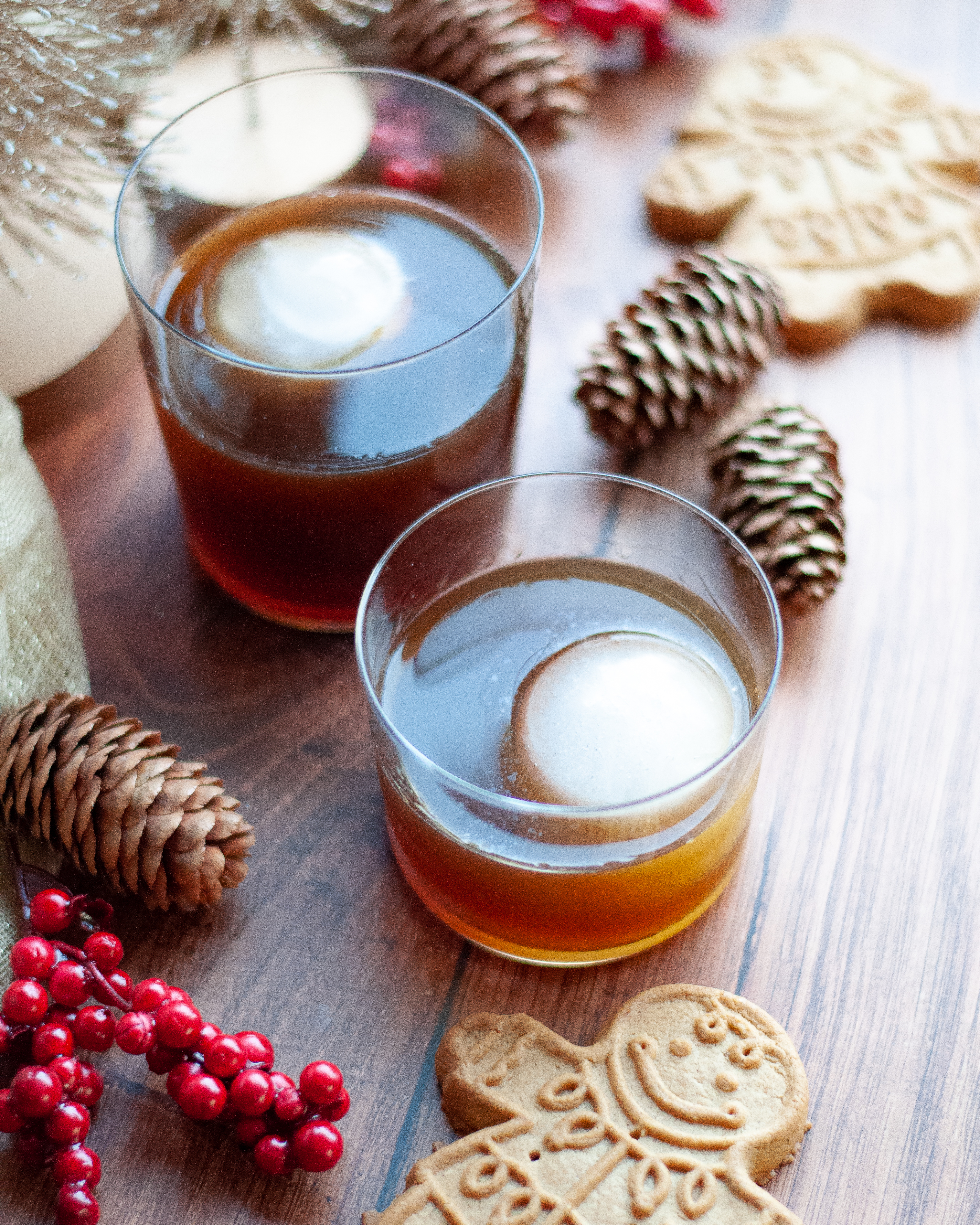 Two gingerbread old fashioneds with ice spheres ready to be drank. The Christmas old fashiond glasses are surrounded by smiling gingerbread cookies, pinecones, red berries, and gold trees.