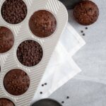 a muffin tin filled with alternating double chocolate zucchini muffins and chocolate chips. additional muffins and chocolate chips sit outside of the pan.
