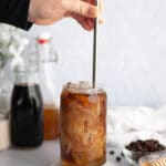 Image of a glass of honey almondmilk cold brew being stirred with a metal straw. The glass is sitting on a white coaster, and next to a bowl of honey and bowl of coffee grounds. In the background is a white linen, and glass containers of honey simple syrup, cold brew coffee, almond milk.