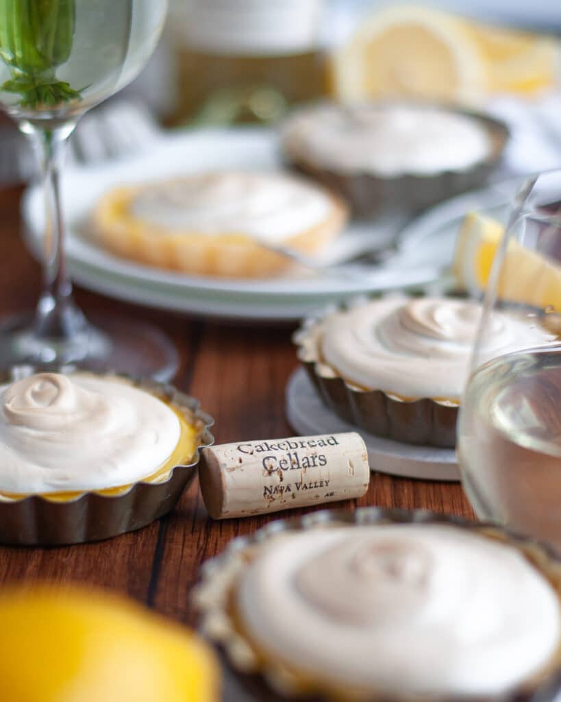 Many lemon meringue tartlets on display surrounded by tart pans, lemon slices, and wine glasses. A wine cork is in focus in the center of the image.