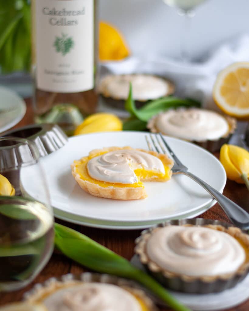 Close up of a lemon meringue tartlet on a white plate with a slice taken out of it. The plate is surrounded by tart pans, lemon slices, and yellow tulips.
