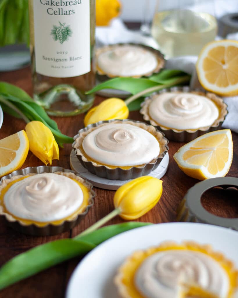 Many lemon meringue tartlets on display among tart pans, yellow tulips, lemon slices, and a bottle of Cakebread Sauvignon Blanc and wine glasses filled with white wine.