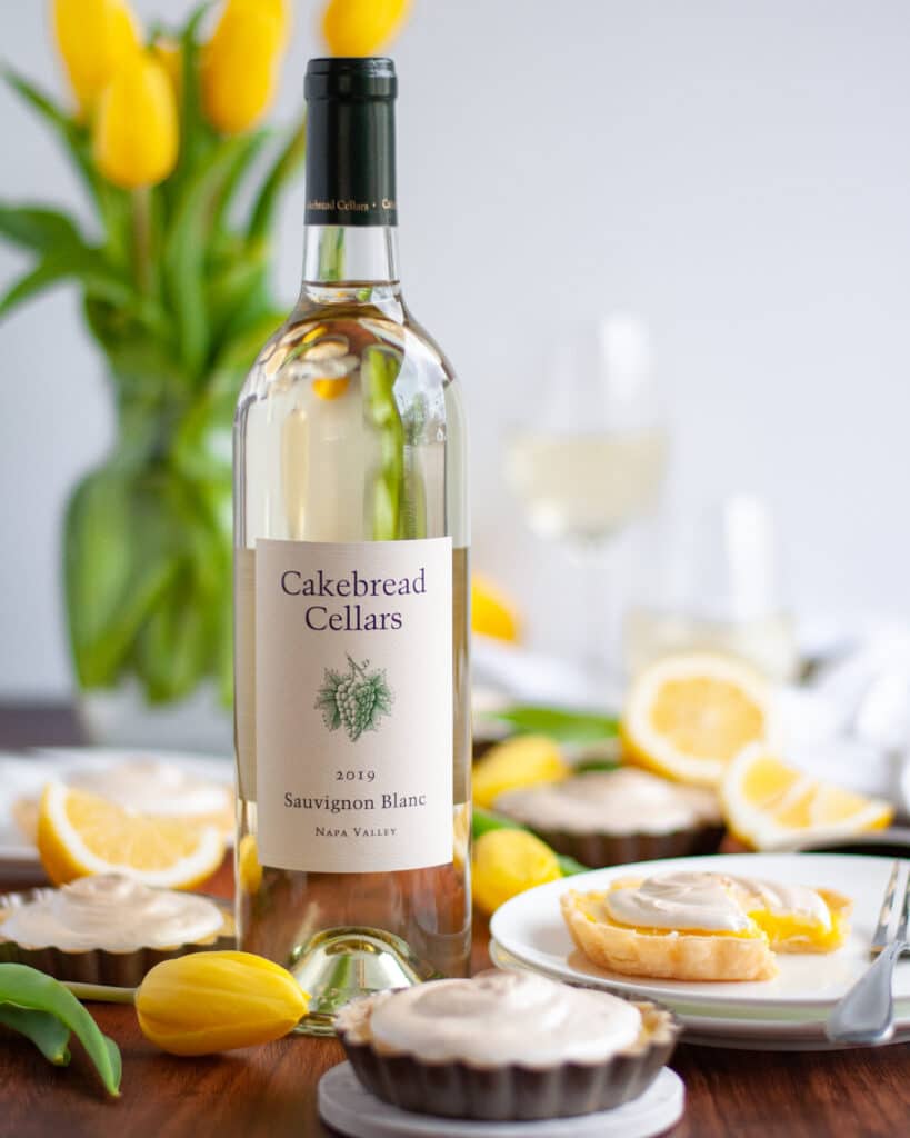 A bottle of Cakebread Sauvignon Blanc wine surrounded by lemon tarts, lemon slices, yellow tulips, and glasses of wine.