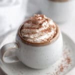 close up of a mug filled with homemade hot chocolate and topped with whipped cream and chocolate shavings. The mug sits on a plate and is surrounded by pieces of chocolate and another mug of hot chocolate in the background.