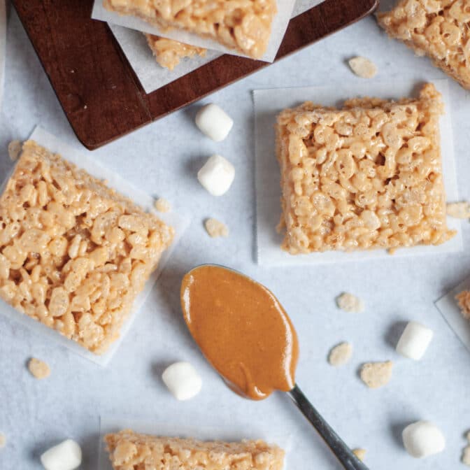 top down view of several peanut butter rice krispy squares. surrounded by mini marshmallows, rice krispie cereal, and a spoon full of creamy peanut butter