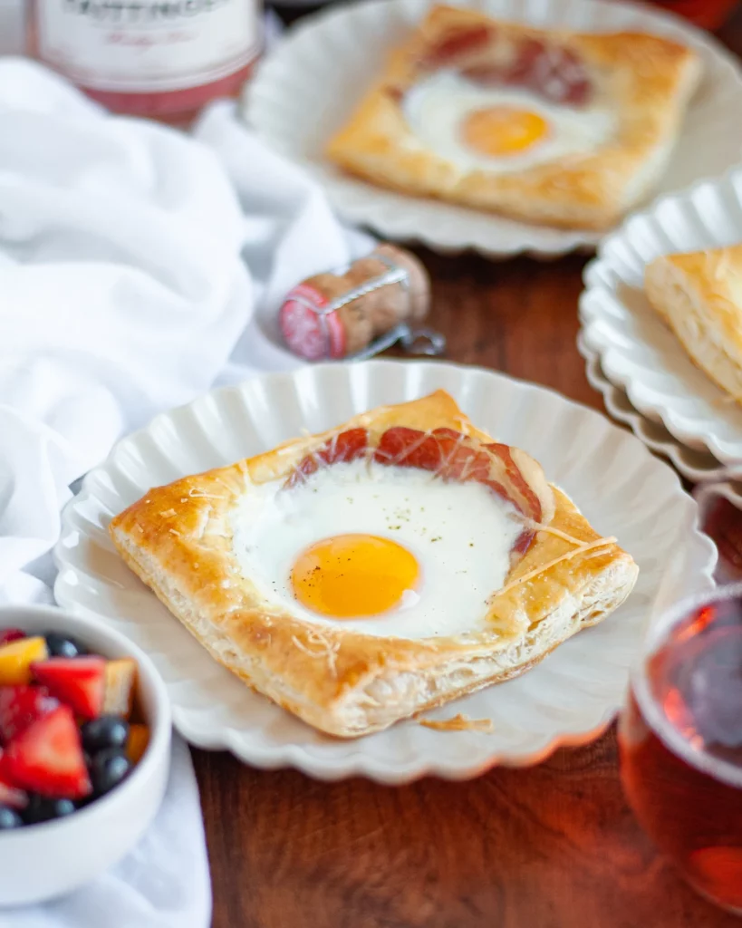 A puff pastry egg tart on a cream plate with ruffled edges. The main tart sits near to a glass of pink champagne, a small bowl of fruit salad, a white linen, and a champagne cork. Two additional plates hold egg tarts with puff pastry are in the background as well.