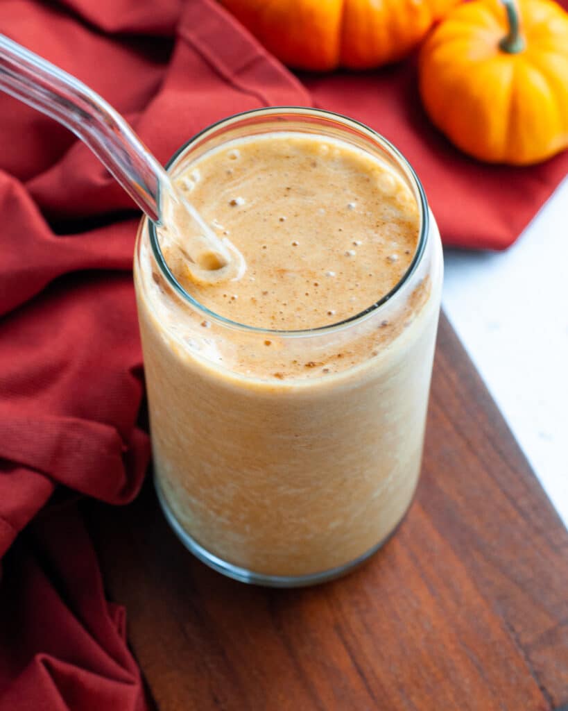 Close up of a pumpkin pie protein shake with a glass straw. The protein shake is sitting on a wooden cutting board, and is next to an orange napkin and pumpkins.