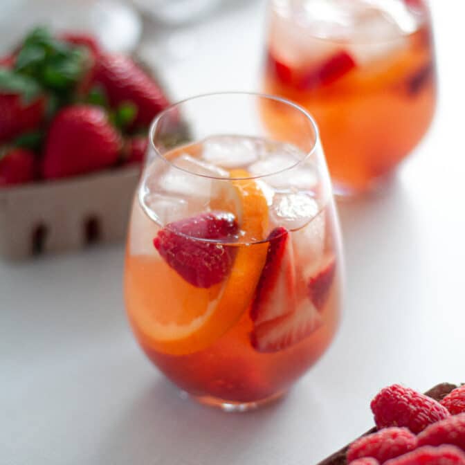 close up of a stemless wine glass filled with rose berry sangria. additional glasses are in the background, and cardboard containers of fresh berries surround the glass.