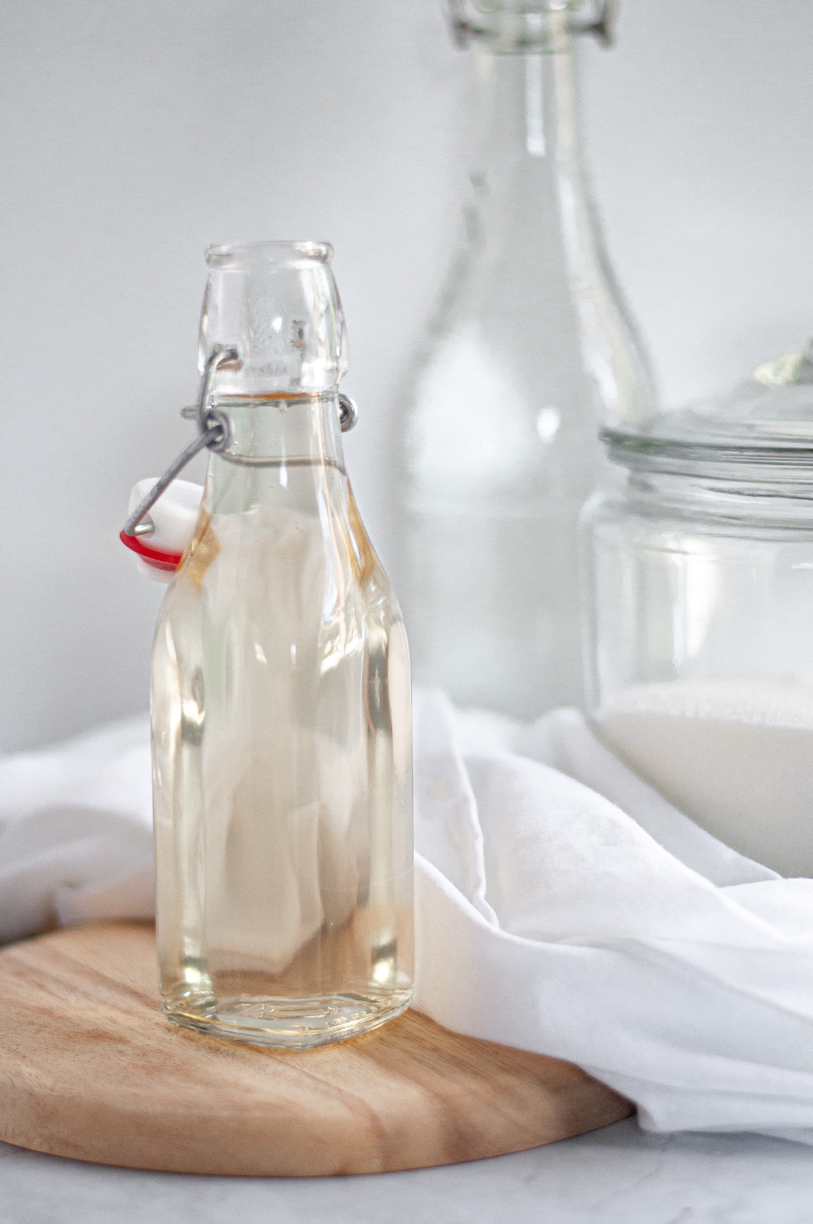 Container of simple syrup on a wooden block, with a napkin, jug of water, and container of sugar in the background.