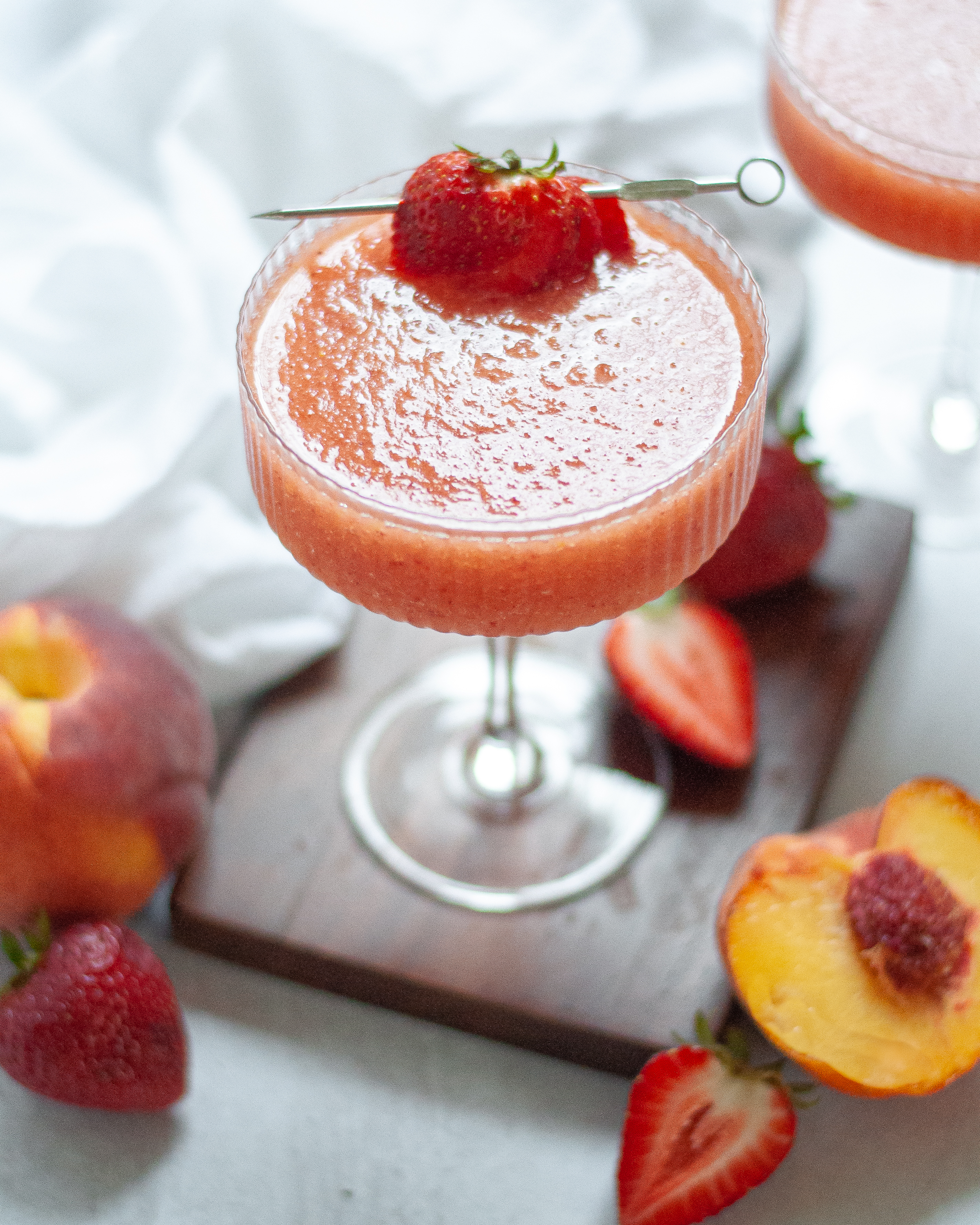 Coupe glass filled with this strawberry peach frose recipe. The glass is garnished with a sliced strawberry on a cocktail stick and it is sitting on a dark wooden board. The wine cocktail is surrounded by fresh fruit, a white napkin, and another cocktail is peaking into the top corner of the picture.