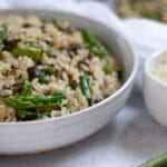 Close up of a serving bowl of this lemon risotto with asparagus and kale. There is fresh asparagus in the foreground, and a cup of grated parmesan and two plates with risotto on it in the background.