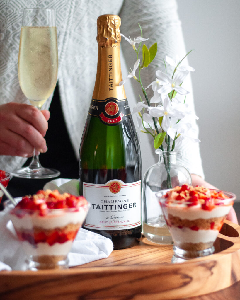 A tray with a bottle of Taittinger Brut Champagne, no bake cheesecake cups, flowers, and a linen. A person is behind the tray, holding a glass of champagne, and looks to be about to pick up the tray.