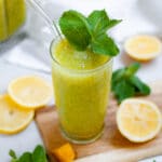 close up of a tall glass filled with mango mint lemonade with a glass straw an mint leaf garnish. the glass is is surrounded by lemon slices, mango chunks, and leaves of fresh mint and has the blender of mango mint puree in the background.