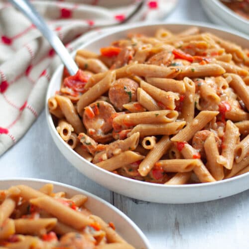 close up of a serving bowl filled with this chicken andouille sausage cajun pasta. plates filled with pasta surround the serving dish as well as a red and tan linen.